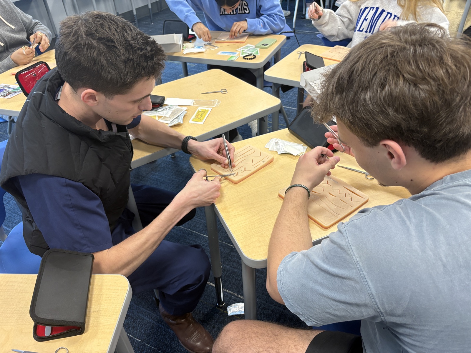 Two students practicing sutures on training pads with surgical instruments